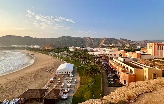 منظر لشاطئ بندر الجصه في مسقط، مع الرمال الذهبية، والمباني الفاخرة، والجبال الشاهقة في الخلفية.